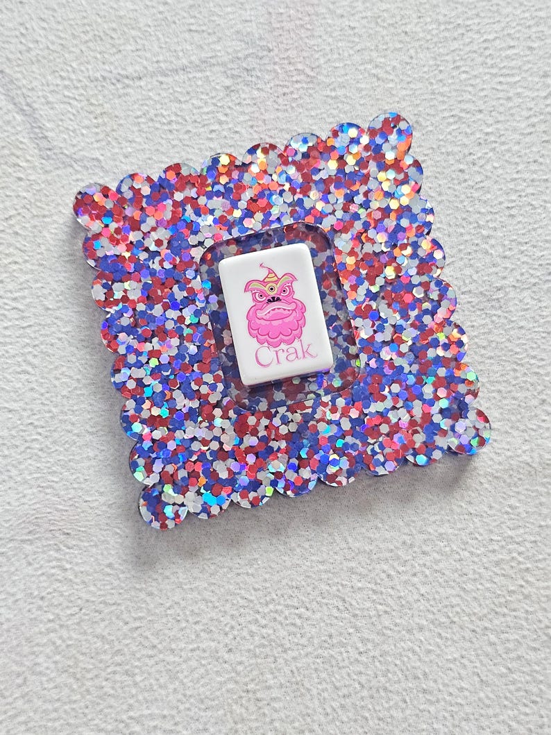 Peut inclure: Un dessous de verre carr&eacute; et scintillant avec un bord festonn&eacute;. Le dessous de verre est rempli de paillettes rouges, blanches et bleues. Un insert rectangulaire blanc au centre pr&eacute;sente un personnage de dessin anim&eacute; rose et le mot "Crak".