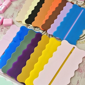 May include: A collection of colorful, wavy-edged silicone game tile holders in various colors, including pink, blue, green, and yellow. The holders are designed to hold game tiles, with a slot for easy access. Pink game tiles are scattered around the holders.