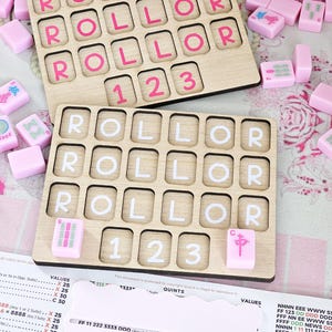 May include: Two rectangular wooden game boards with grid cutouts, each featuring the word ROLLOR and the numbers 1 2 3. The top board has pink lettering, while the bottom board has white lettering. Scattered pink mahjong tiles surround the boards.