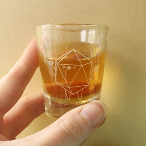 May include: A clear glass shot glass with a geometric design of a cartoon character holding a small glass. The glass is filled with a light amber liquid.