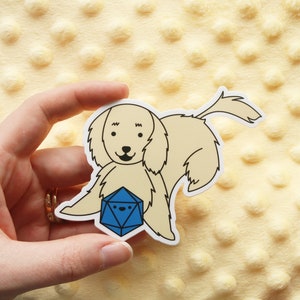 May include: A white dog with brown eyes and a brown nose is holding a blue and white dice with a smiley face. The dog is sitting on a yellow surface.