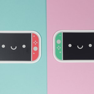 May include: Two Nintendo Switch game console stickers with a cute cartoon face. The sticker on the left has a blue and red color scheme, while the sticker on the right has a green and pink color scheme.