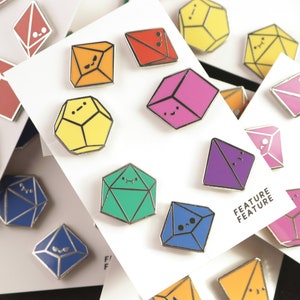 May include: A set of colorful enamel pins shaped like geometric dice with smiling faces. The pins are arranged on white cards with the text "Feature Feature" printed on one of the cards.