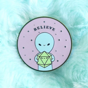 Peut inclure: Une épingle en émail rose et or avec un extraterrestre bleu tenant un dé vert à plusieurs faces. L'épingle indique "Believe" en lettres blanches.