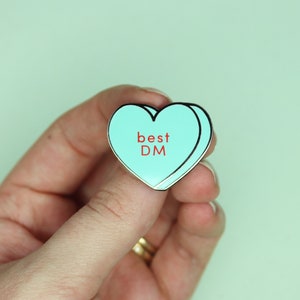 May include: A heart-shaped enamel pin in light blue with a black outline. The pin features the words "best DM" in red lettering. The pin is held in a hand against a light green background.