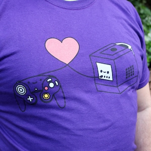 May include: Purple t-shirt featuring a graphic of a video game controller and console connected by a pink heart. The controller and console have cartoon faces. The shirt is a solid purple color.