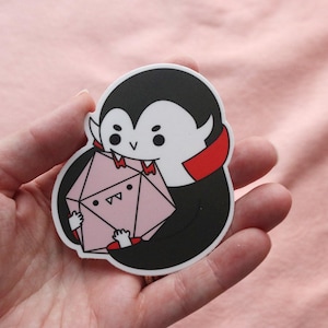 May include: A sticker featuring a cartoon vampire with a black cape and red collar, holding a pink, geometric die. The vampire has a white face and is smiling. The sticker is held in a person's hand against a pink background.