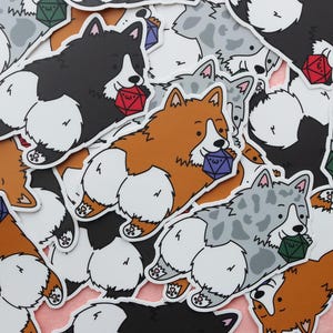 Puede incluir: Una colección de pegatinas de corgi en varios colores, incluyendo negro, marrón y gris. Cada pegatina presenta un corgi sosteniendo un dado multicolor, lo que sugiere un tema relacionado con los juegos de mesa o juegos de rol.