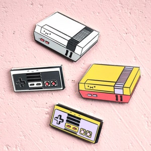 May include: Four enamel pins in the shape of retro video game consoles and controllers. The pins feature a white, yellow, and black color scheme with red accents. The pins are arranged on a pink textured surface.