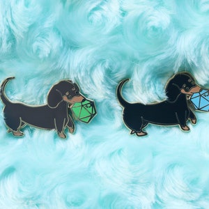 May include: Two black dachshund pins with gold accents. One dachshund is holding a green die and the other is holding a blue die. The pins are on a light blue fuzzy background.