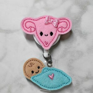 May include: A badge reel featuring a pink felt uterus shape with a small pink bow and a smiling face, attached to a dangling felt baby wrapped in a blue blanket with a pink heart, featuring a tan face and a small swirl of hair on the head.