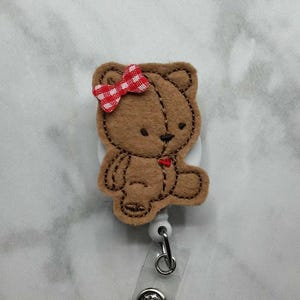 May include: A brown teddy bear badge reel with a red and white gingham bow. The bear is made of felt and has a stitched outline. The reel has a clear plastic strap and a metal clip.