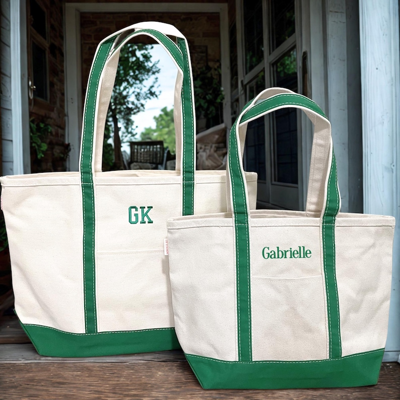 May include: Two canvas tote bags with green trim. The bags have cream-coloured bodies, green bases, and green straps. One bag is embroidered with the initials "GK", the other says "Gabrielle".
