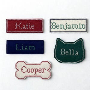 May include: Five felt name tags with cross-stitch embroidery. A red rectangle with 'Katie', a white rectangle with 'Benjamin', a blue rectangle with 'Liam', a dark green cat-shaped tag with 'Bella', and a white bone-shaped tag with 'Cooper'.