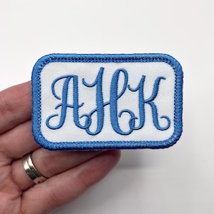 May include: Embroidered patch with the letters "AHK" in a blue script font on a white background. The patch has a blue border and is rectangular in shape. The patch is likely for clothing or accessories.