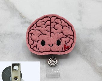 May include: A pink felt brain badge reel featuring dark pink embroidered brain folds, black eyes, a small smile, and a red heart accent. The badge reel includes a silver metal clip and a clear plastic strap, with a cordless badge clip shown.