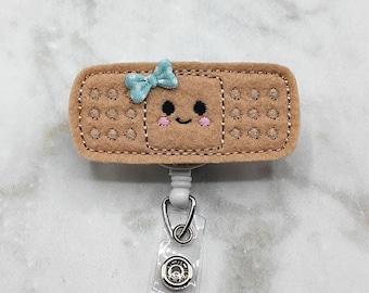 Puede incluir: Un carrete de insignia de fieltro marrón con un diseño de vendaje sonriente con un lazo azul claro. El carrete de la insignia tiene una correa de plástico transparente y un clip de metal. El vendaje tiene detalles bordados y una cara linda.
