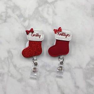 May include: Two badge reels featuring red Christmas stocking designs with white cuffs. One stocking has a red glitter finish and the name Emily, while the other is solid red with the name Kaitlyn. Both have small red bows and clear plastic straps.