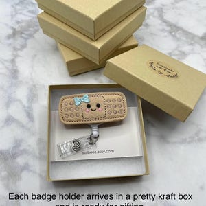A handmade badge holder with a smiley face and a clear plastic keychain attached to it, placed in a cardboard box.