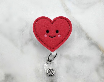 May include: A red felt heart badge reel featuring an embroidered smiling face with black eyes, a curved mouth, and small pink cheeks. The heart is stitched along the edges and attached to a clear plastic retractable cord and a metal clip.