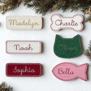 May include: A collection of personalized felt name tags in various shapes and colors. The tags include rectangular, bone-shaped, cat-shaped, and fish-shaped designs, each embroidered with a different name in cursive script. The color palette includes white, burgundy, green, and pink.