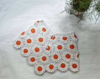 Op de afbeelding: Witte gehaakte rok met oranje margrietvormige bloemen. De rok heeft een gegolfde zoom en een strik aan de taille.