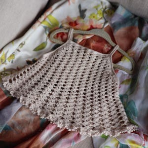 Peut inclure: Un haut en crochet beige avec un ourlet festonné et de fines bretelles, présenté sur un cintre à pois. Le motif ajouré est visible. Le haut repose sur un tissu à motifs floraux.