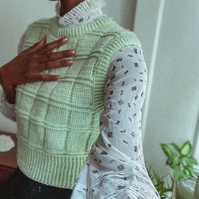 May include: A light green crocheted vest with a square pattern. The vest is worn over a white blouse with a floral print.