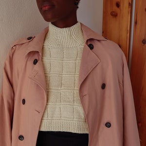 Può includere: Un trench rosa chiaro con un maglione bianco a collo alto di lana sotto. Il maglione ha un motivo a griglia.