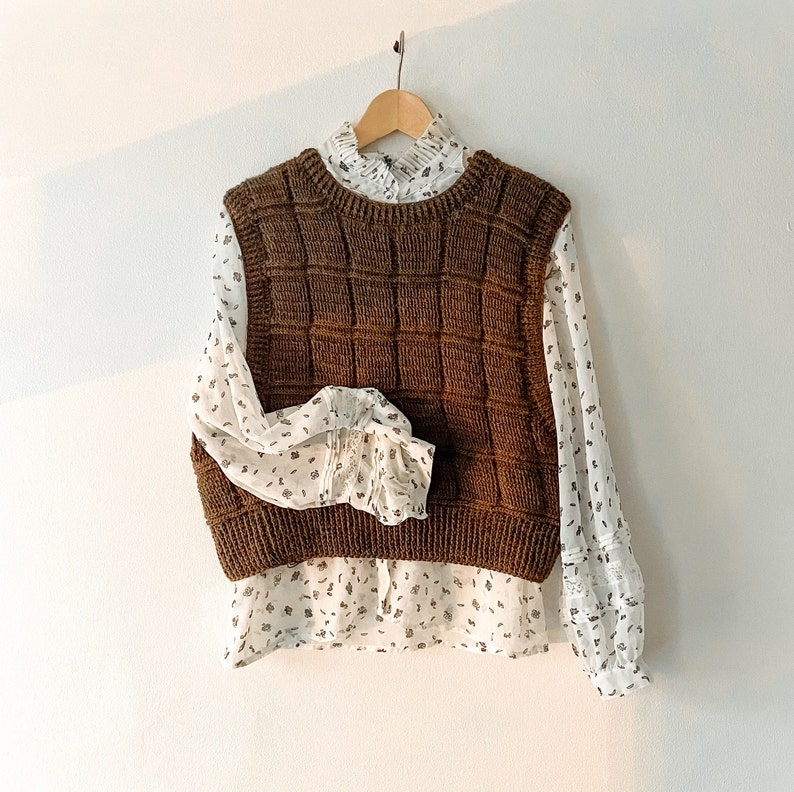 May include: A brown knitted vest with a square pattern, layered over a white blouse with a floral print.