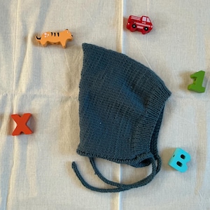 May include: A teal knitted baby bonnet with tie strings, surrounded by wooden toy blocks shaped like a tiger, a fire engine, the letter X, the number 1, and the letter B. The bonnet is made of a textured knit fabric.
