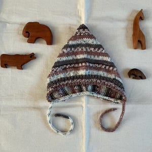 May include: A knitted baby hat in brown, grey, and blue stripes, with ear flaps and ties. Wooden animal toys, including a bear, cow, giraffe, and a small bear, are arranged around the hat.
