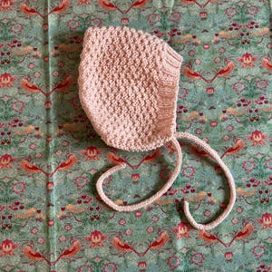 May include: A pale pink knitted baby bonnet with ties, set against a patterned teal fabric with floral and bird motifs. The bonnet is made of a textured knit and has a rounded shape.