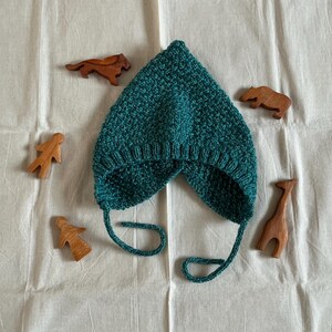 May include: A teal knitted baby hat with ear flaps and ties, alongside small wooden animal figurines. The hat features a textured stitch. The wooden animals include a tiger, giraffe, rhino, and human figures. The items are arranged on a white fabric surface.