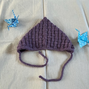 May include: A knitted purple baby bonnet with a textured pattern and attached ties. Two origami paper cranes in blue with white floral patterns are placed on either side of the bonnet. A charming scene.
