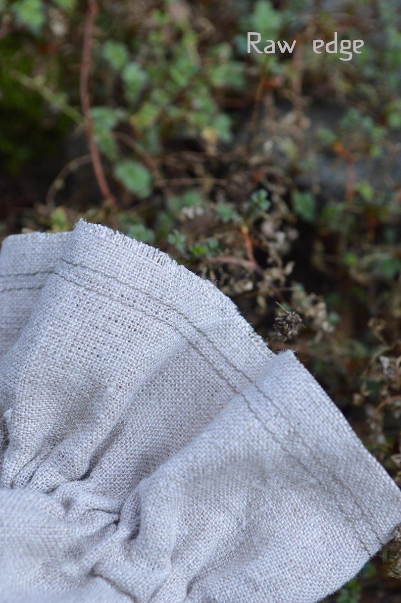 May include: Close-up of folded, light beige linen fabric with a raw edge. The fabric is textured and has visible stitching along the edge. The words "Raw edge" are in the top right corner.
