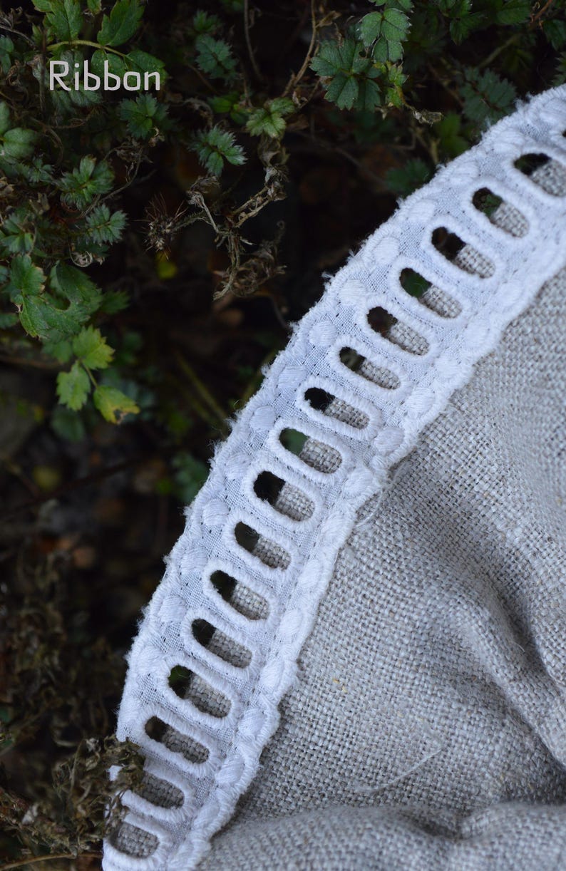 May include: Close-up of a white ribbon with a scalloped edge, featuring a series of oval cutouts. The ribbon is sewn onto a light grey fabric. The word "Ribbon" is visible in the upper left corner.