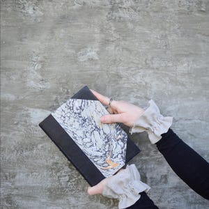 May include: A black journal with a marbled cover design in shades of white, grey, and orange. The journal is held against a grey background.