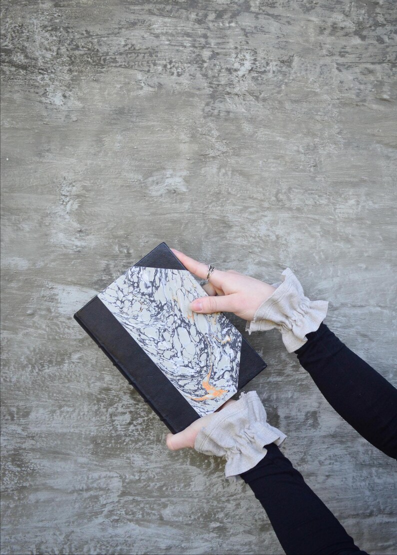 May include: A black journal with a marbled cover design in shades of white, grey, and orange. The journal is held against a grey background.