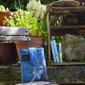 Op de afbeelding: Een blauwe denim boekenhoes met een knoopsluiting, gemaakt van gerecyclede jeans. De boekenhoes staat op een houten bank in een tuin, met planten en boeken op de achtergrond.