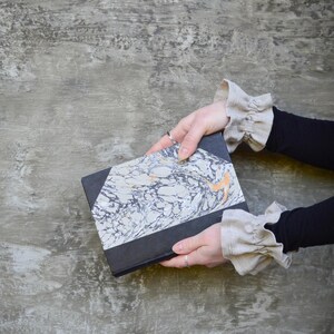 May include: A hardback book with a black spine and a marbled cover in shades of grey, white, and orange. The book is held against a grey textured background.