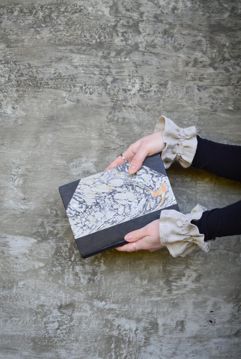 May include: A hardback book with a black spine and a marbled cover in shades of grey, white, and orange. The book is held against a grey textured background.