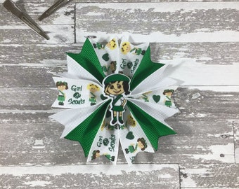 May include: Green and white ribbon hair bow with a girl scout design. The bow features a cartoon girl scout with the words "Girl Scouts" on it.