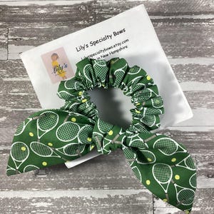 May include: A green scrunchie with a tennis racket and ball pattern. The scrunchie has a bow detail and is on top of a white paper with the text "Lily's Specialty Bows". The scrunchie is a hair accessory.