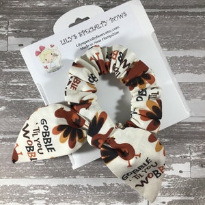 May include: A white scrunchie with a brown and orange turkey print. The scrunchie has the text "Gobble 'Til You Wobble" printed on it.