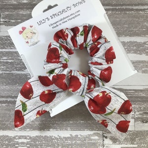 May include: A white and red apple print scrunchie with a bow. The scrunchie is made of fabric and has a white background with red apples printed on it. The scrunchie is tied in a bow and is ready to be worn.