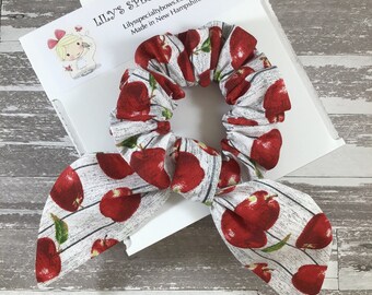 May include: A white and red apple print scrunchie with a bow. The scrunchie is made of fabric and has a white background with red apples printed on it. The scrunchie is tied in a bow and is ready to be worn.