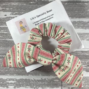 May include: A green, red, and white patterned scrunchie with a bow. The scrunchie has a candy cane and heart design. The scrunchie is on a white surface with the text "Lily's Specialty Bows" and "Lilyspecialtybows.etsy.com Made in New Hampshire".