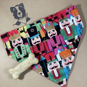 May include: A triangular dog bandana with a festive Christmas nutcracker pattern. The bandana is red, green, blue, and black with a white background. The nutcrackers are wearing colorful hats and have white faces with black mustaches.