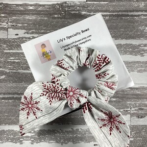May include: A white and gray scrunchie with a red snowflake pattern. The scrunchie is tied in a bow and is on a white background. Lily's Specialty Bows, Lilyspecialtybows.etsy.com, Made in New Hampshire.